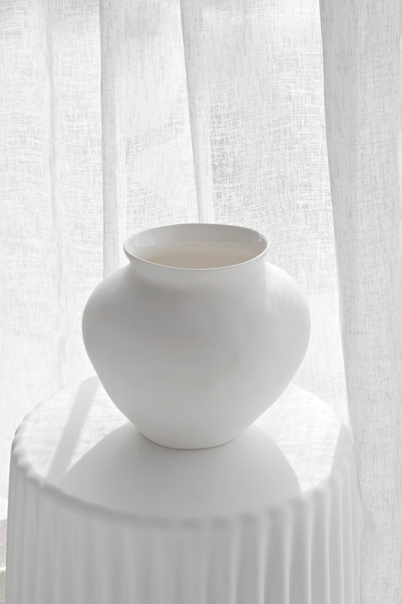 Venus Vase in matte white ceramic styled on a pedestal with soft natural light filtering through sheer curtains.