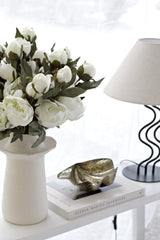 Lifelike white peony stems arranged in a neutral ceramic vase beside a lamp and coffee table books in soft natural light.