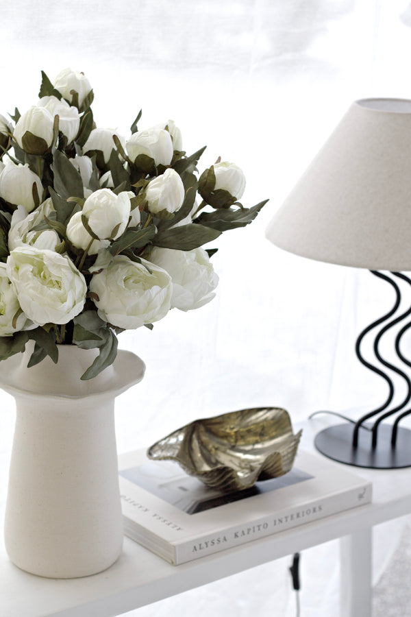 Lifelike white peony stems arranged in a neutral ceramic vase beside a lamp and coffee table books in soft natural light.