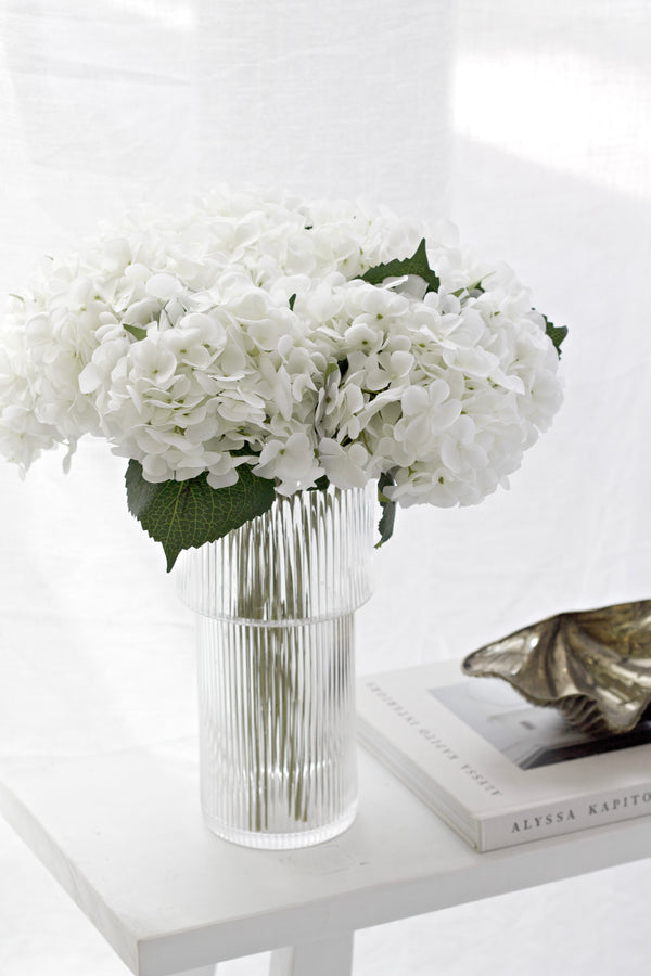 White artificial hydrangea stems in ribbed glass vase on white table, styled with books and decor in soft natural lighting