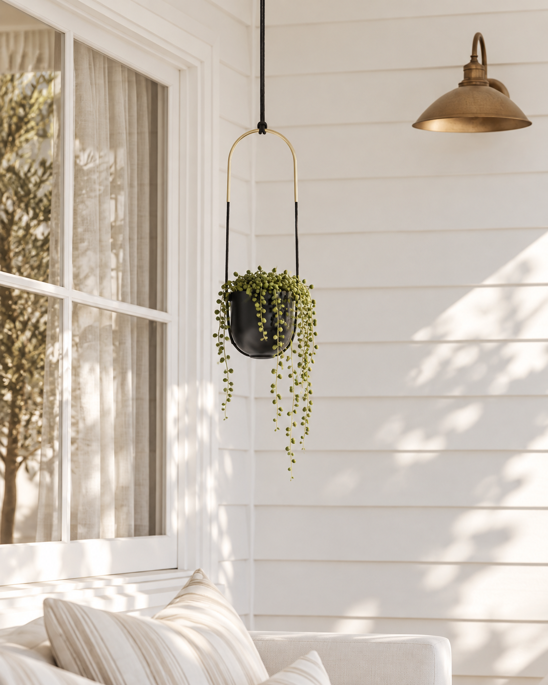 Umbra Bolo hanging planter in black and brass with trailing string of pearls on a white weatherboard porch beside a window