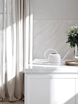 A minimalist white galvanised steel watering can with a long spout styled on a marble kitchen bench next to fresh greenery and neutral curtains.