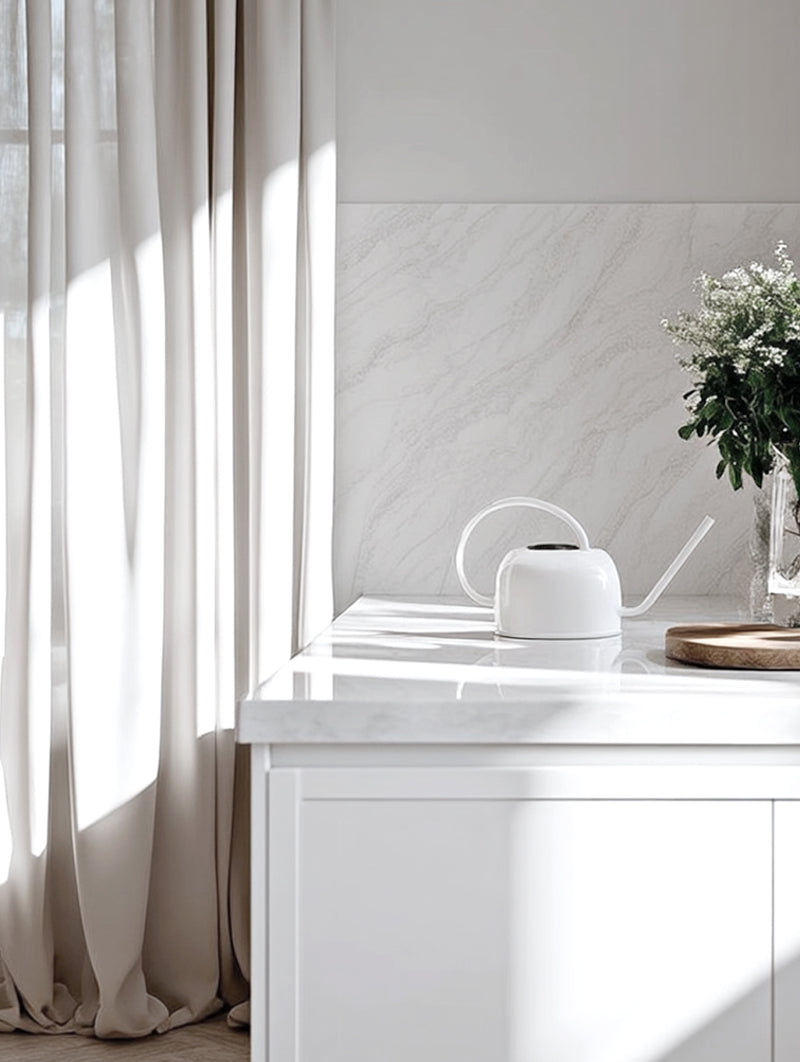 A minimalist white galvanised steel watering can with a long spout styled on a marble kitchen bench next to fresh greenery and neutral curtains.