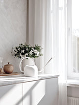 Modern white watering can with a curved handle and long spout styled on a sunny kitchen windowsill beside white curtains, wood accents, and fresh greenery.