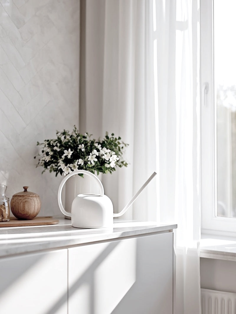 Modern white watering can with a curved handle and long spout styled on a sunny kitchen windowsill beside white curtains, wood accents, and fresh greenery.