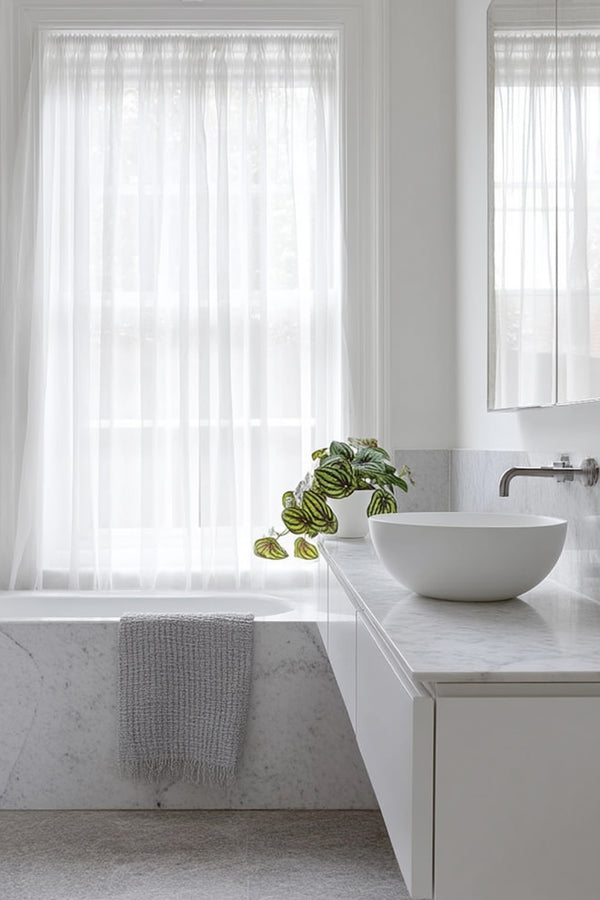 A realistic faux Peperomia Watermelon plant in a white pot placed on a marble bathroom vanity next to a white bowl sink, with soft sheer curtains filtering light.