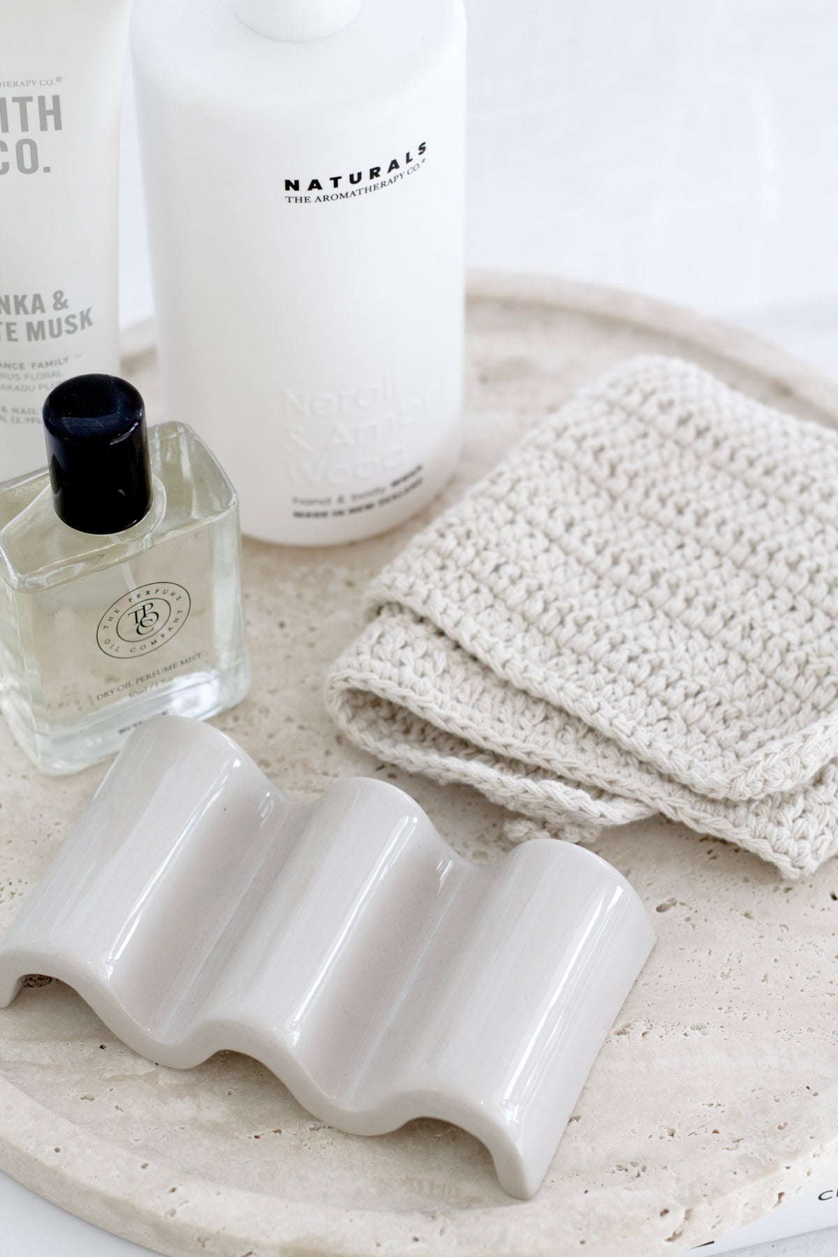 Modern ceramic wave dish styled on a travertine tray with body care products and textured cotton cloth.