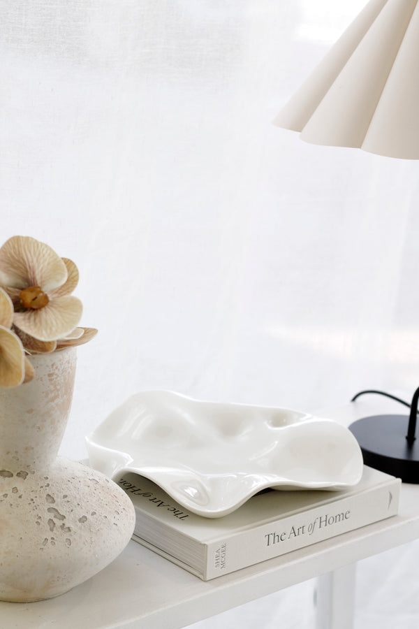 Glossy white wavy ceramic platter styled on a book beside a fluted lamp and sculptural vase with florals in a soft, sunlit room.