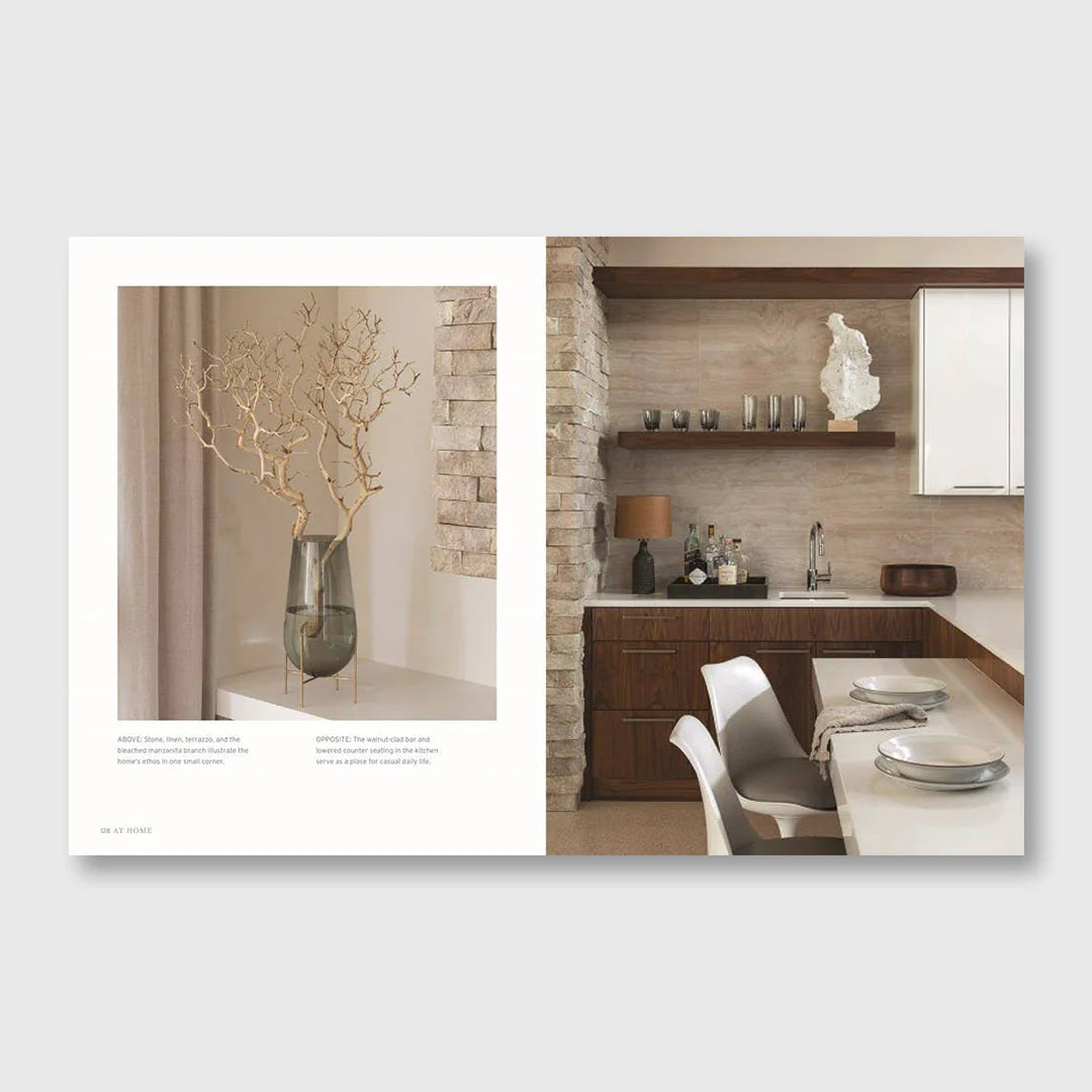 At Home open to a spread of a vase of branches beside a stone-walled kitchen with timber shelves