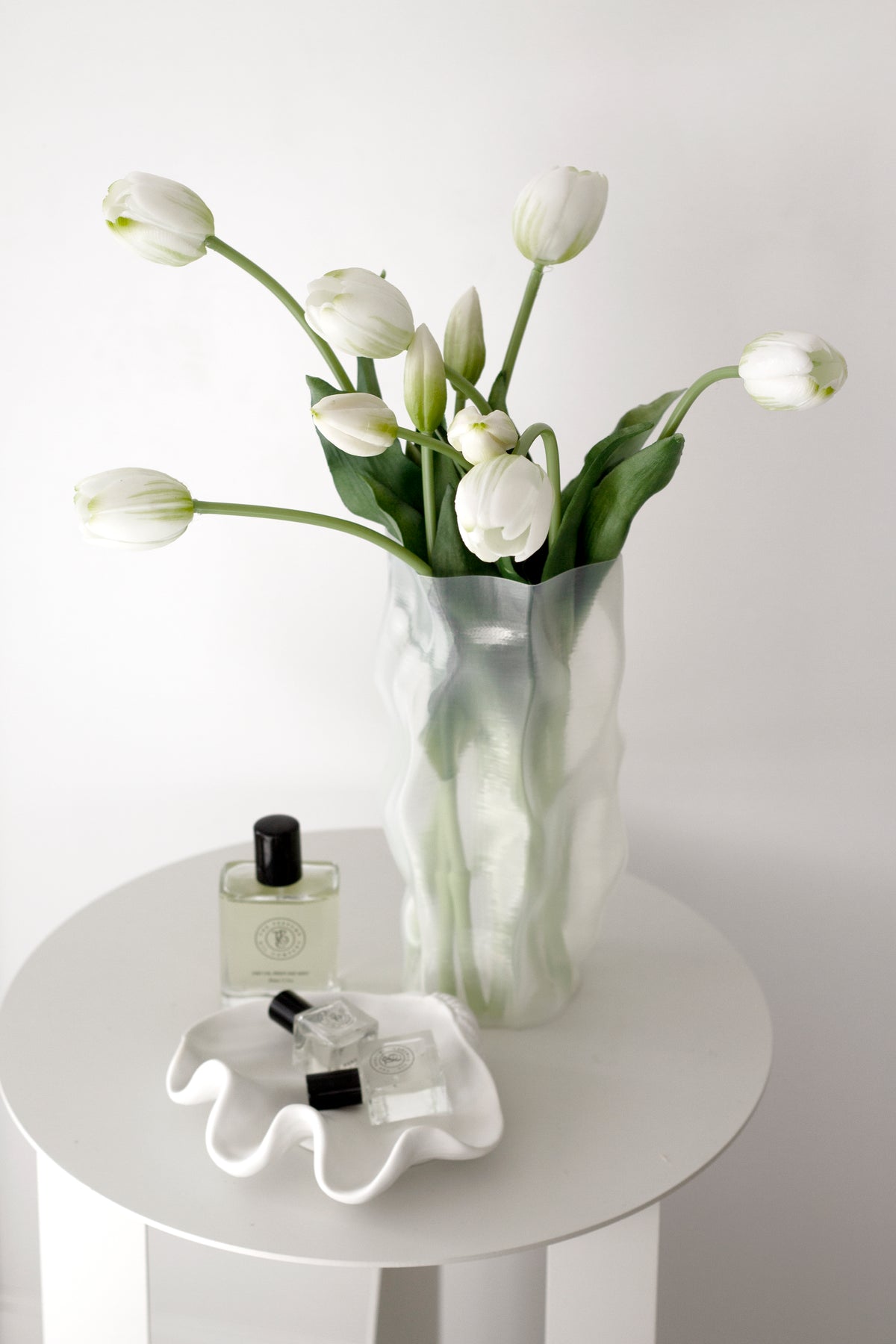 White faux tulip bouquet in a sculptural vase on a white side table, styled with perfume for a calm, minimal look