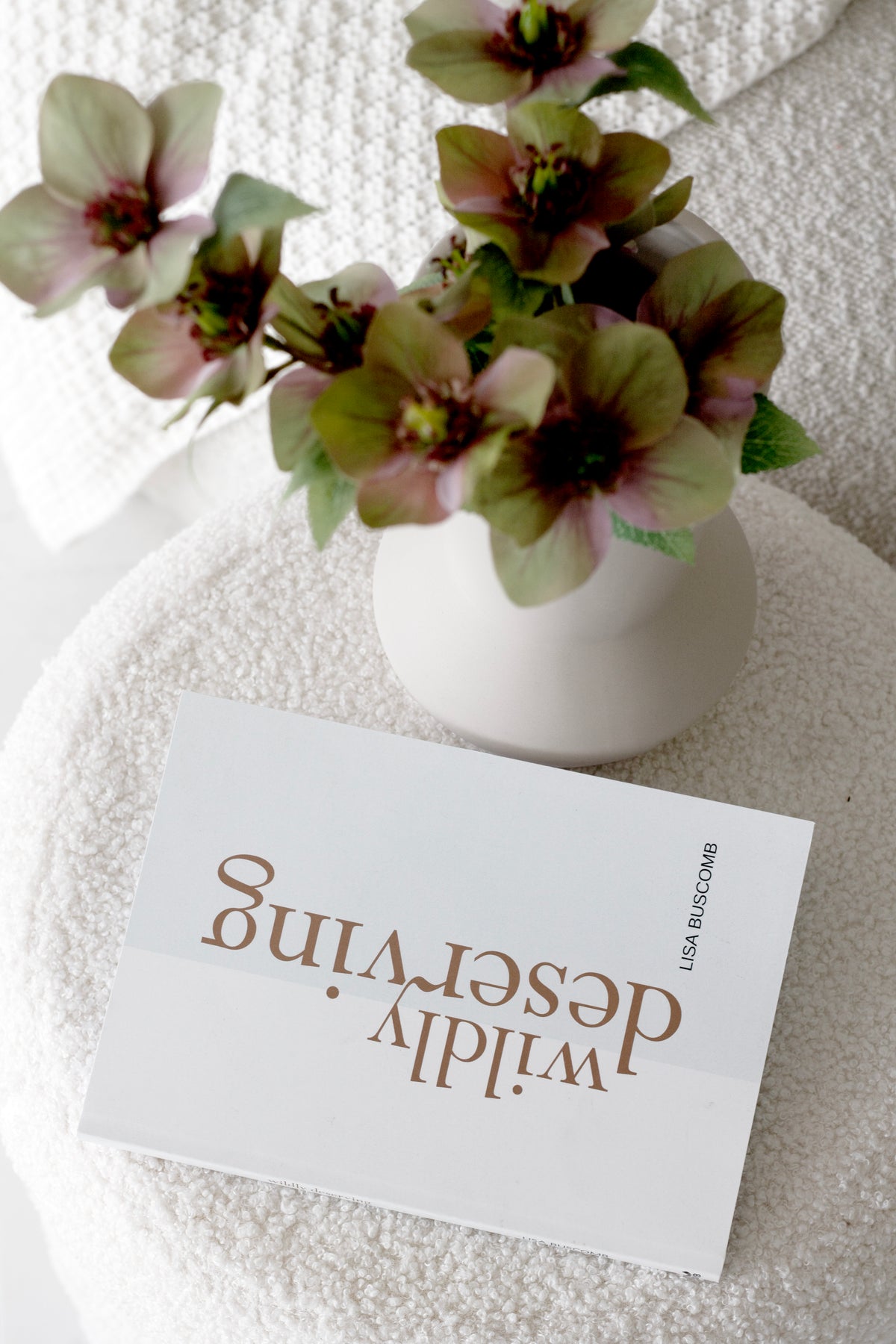 Wildly Deserving book by Lisa Buscomb styled on boucle stool with mauve artificial flowers in ceramic vase – slow living aesthetic