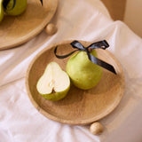 Two green pears on a wooden plate with a black ribbon, one pear is sliced open, on a light fabric background.