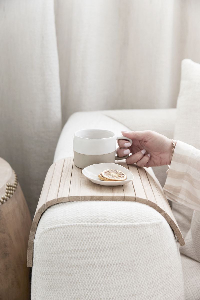 Neutral wooden sofa arm tray holding a ceramic mug and small plate on a cream boucle armchair, with soft curtains behind.