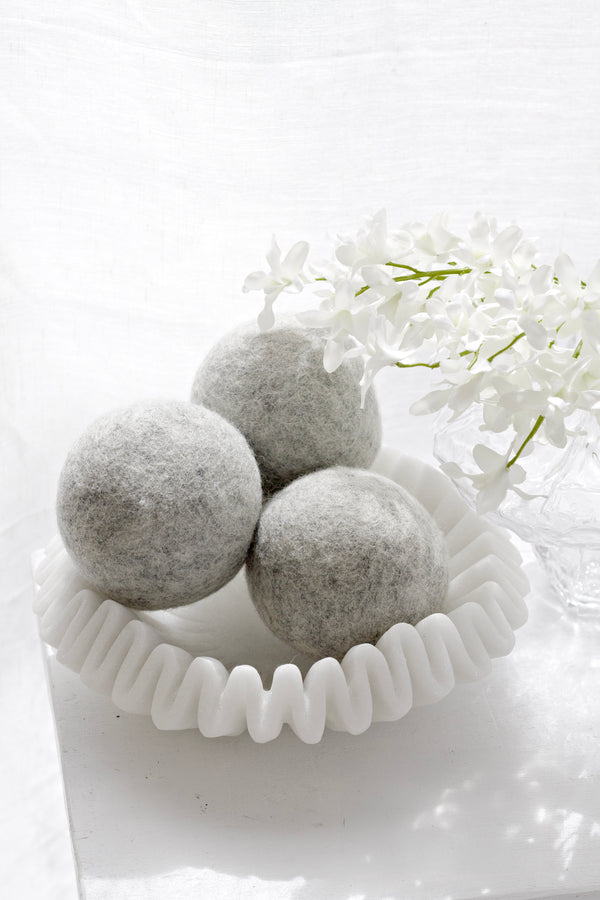 Three grey wool dryer balls displayed in a sculptural white dish with soft white flowers.