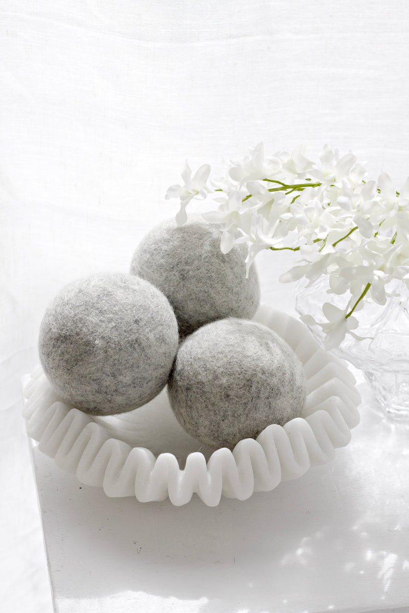 Three grey wool dryer balls displayed in a sculptural white dish with soft white flowers.