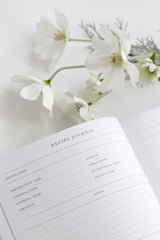 An open Write To Me Recipe Journal showing structured recipe layout pages with fields for recipe name, preparation time, cooking details, notes, and source, styled flat on a white surface with delicate white cosmos flowers scattered softly around it.