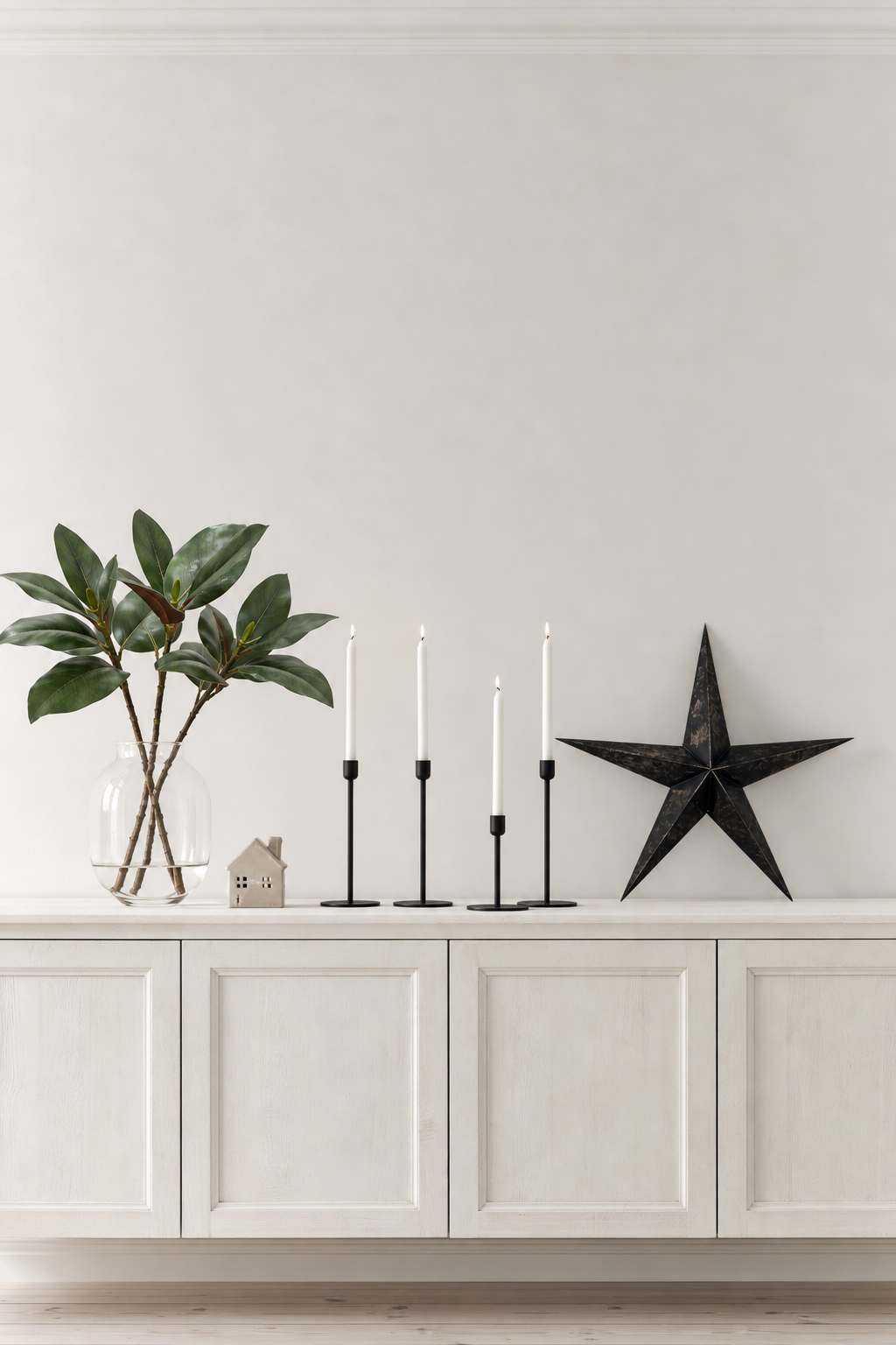 Three Magnolia Leaf Branches in a clear glass vase on a white sideboard beside black candlesticks and a black star