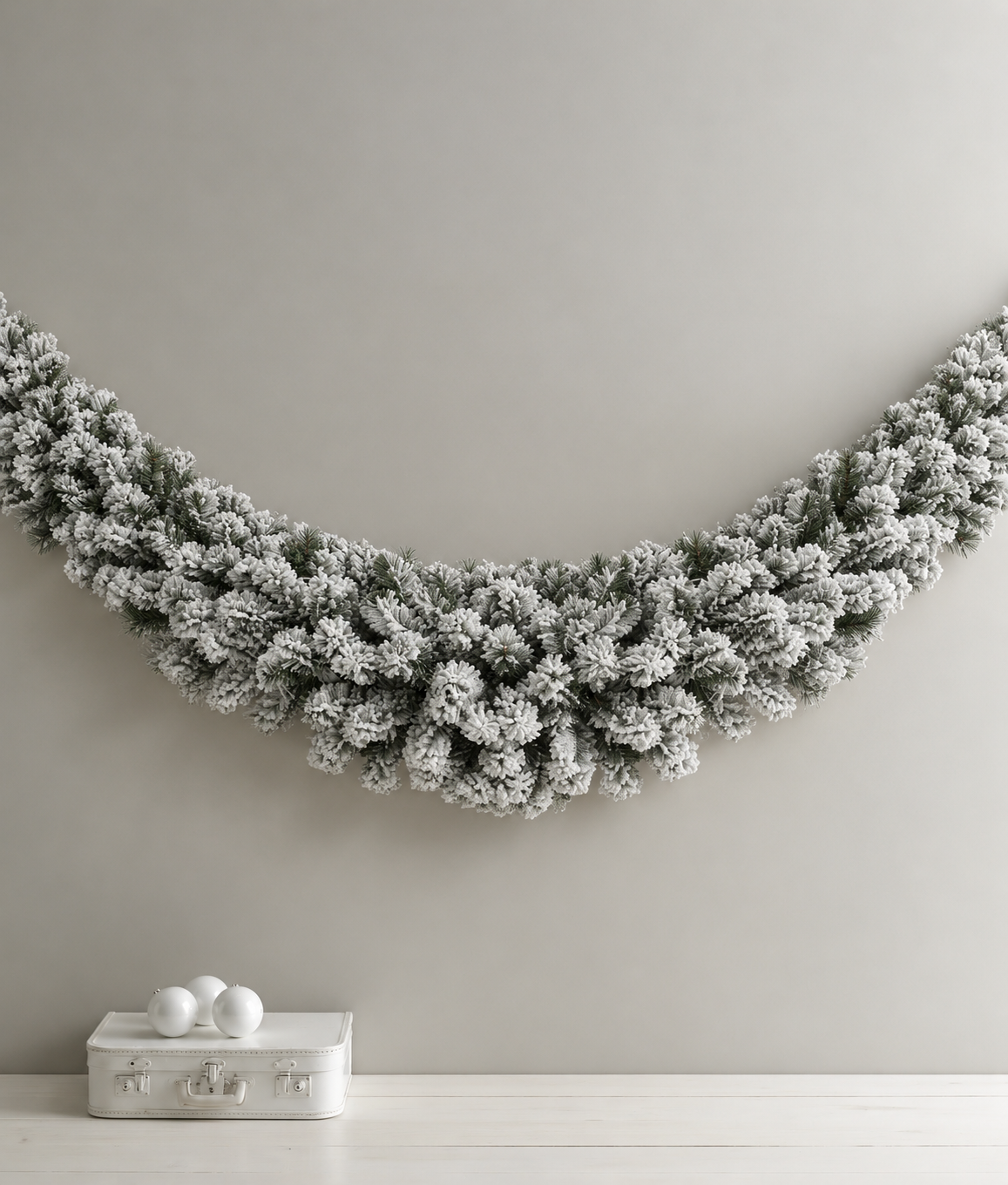 Flocked Royale Christmas Swag hung in a wide curve on a pale grey wall above a white case with baubles