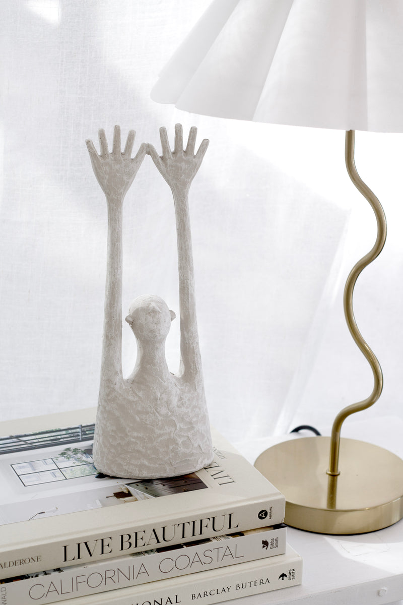 Minimalist white resin figurative sculpture with raised arms, styled on stacked design books beside a brass lamp.