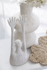 White matte resin Yay figurative sculpture with elongated raised arms styled beside a speckled white ceramic vase with white flowers and woven rattan mat on a white surface