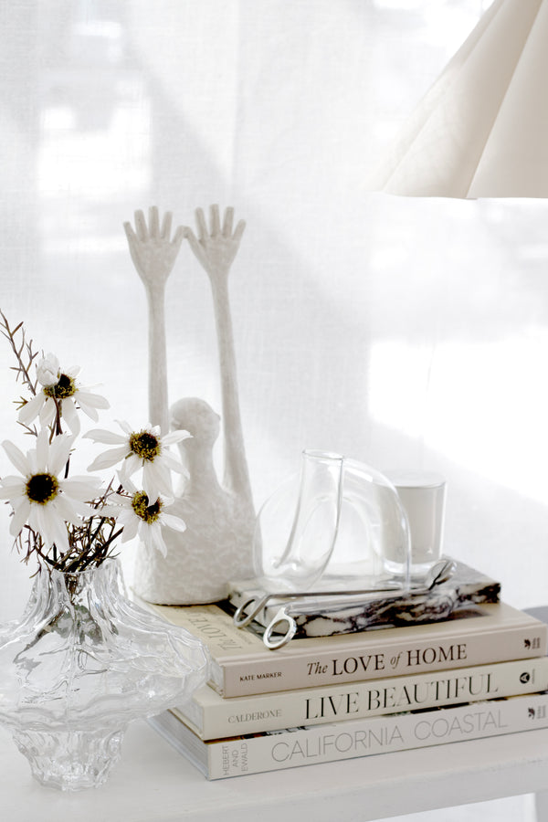 White abstract figurine sculpture with raised arms styled on console table with books, vase, and glass accents.