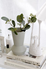 White textured Yay sculpture holding a lampshade, styled beside a matte grey vase with greenery on stacked coffee table books