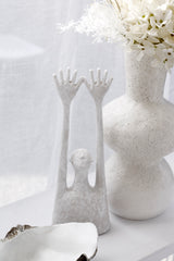 White textured Yay sculpture of a minimal figure with long raised arms beside a speckled white vase of flowers and a white shell bowl on a light surface.