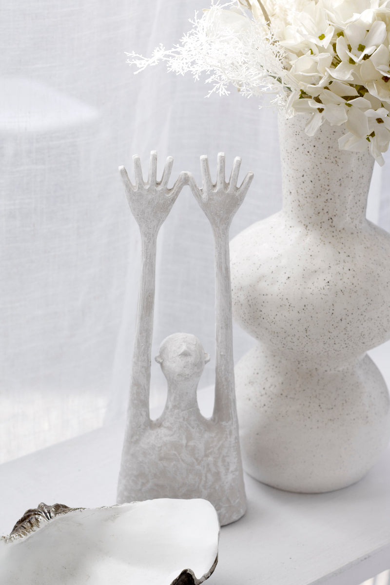 White textured Yay sculpture of a minimal figure with long raised arms beside a speckled white vase of flowers and a white shell bowl on a light surface.