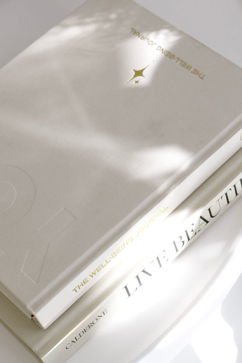 YOU. Wellbeing Journal in sand linen stacked on the book Live Beautiful, in soft natural light on a white surface.