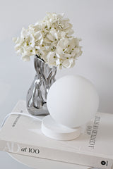Minimalist Yuri Table Lamp with frosted glass globe and powder-coated white steel base, styled on books beside a chrome vase of white flowers