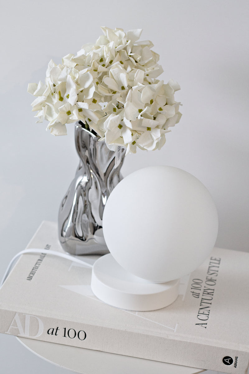 Minimalist Yuri Table Lamp with frosted glass globe and powder-coated white steel base, styled on books beside a chrome vase of white flowers