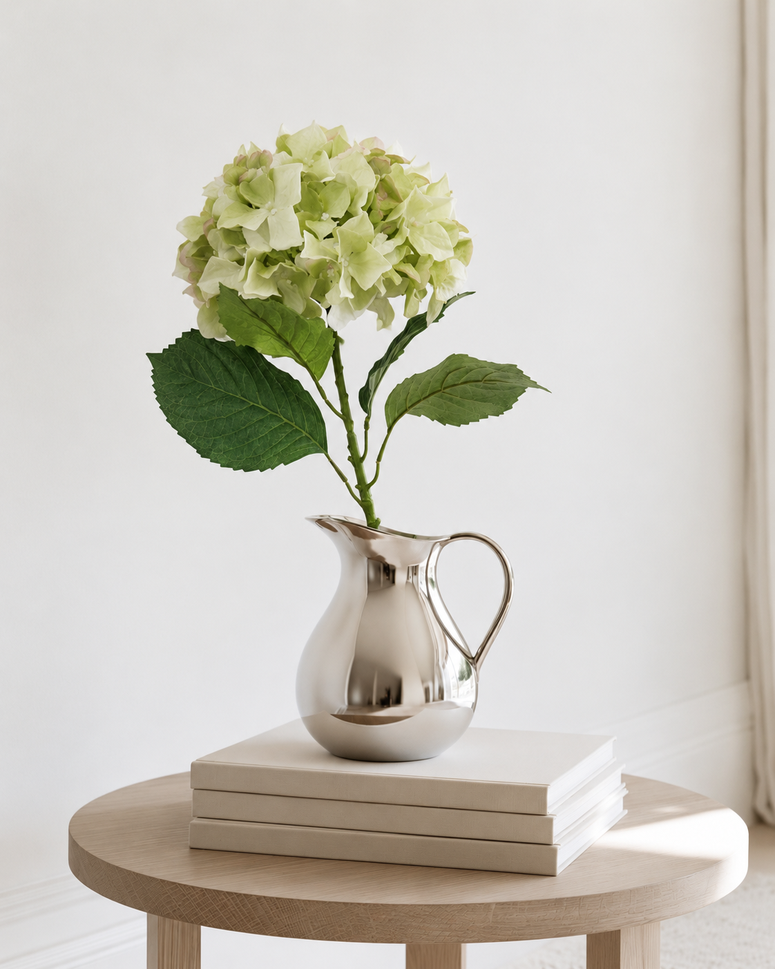 Single green Sublime Hydrangea stem in a silver jug on stacked books on a round oak side table