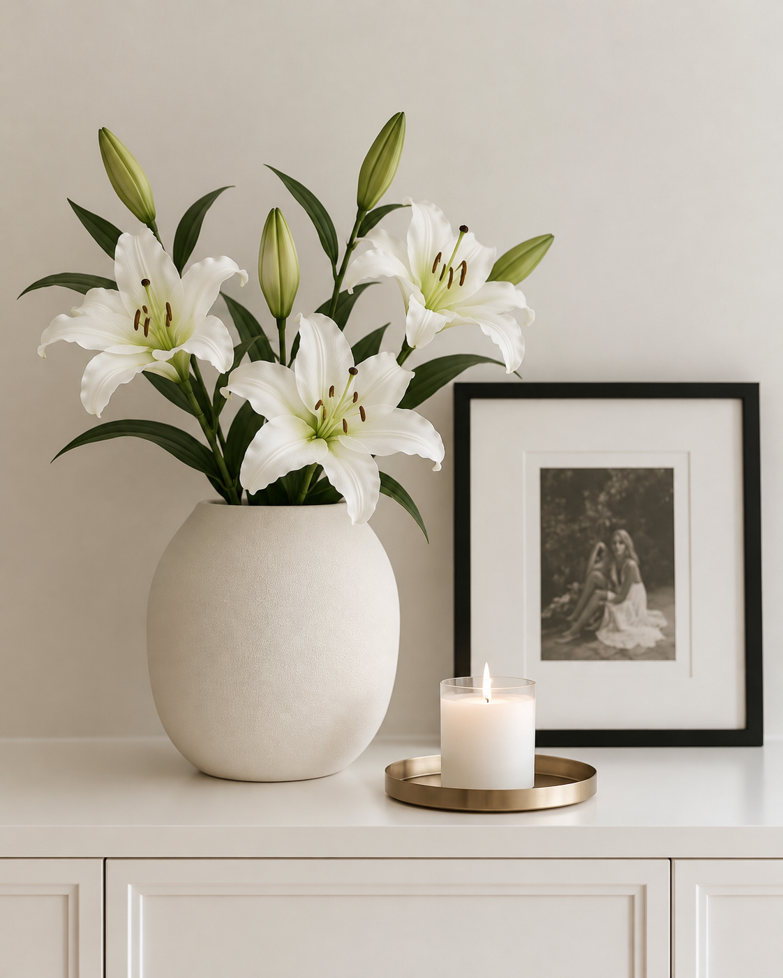 Three white faux rubrum lily stems in a round textured white vase on a white sideboard beside a framed photo and candle