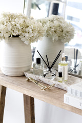 Zakkia terrazzo square tray in seashell tone styled on a timber console with reed diffuser, perfume oils and brass wick trimmer beside large white hydrangeas and a mirror