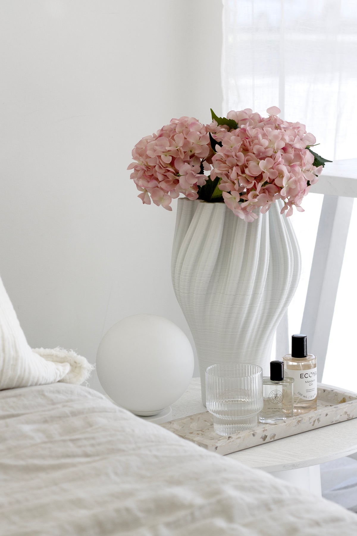 Handmade terrazzo square tray in seashell finish, styled bedside with glassware, perfumes, and pink hydrangeas in a vase.