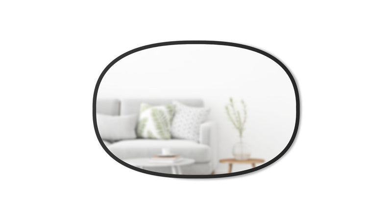 The Hub Mirror Oval - Black from the Umbra range is a stylish addition to any living room. This round mirror, with its sleek rubber rim, effortlessly complements the surrounding couch.