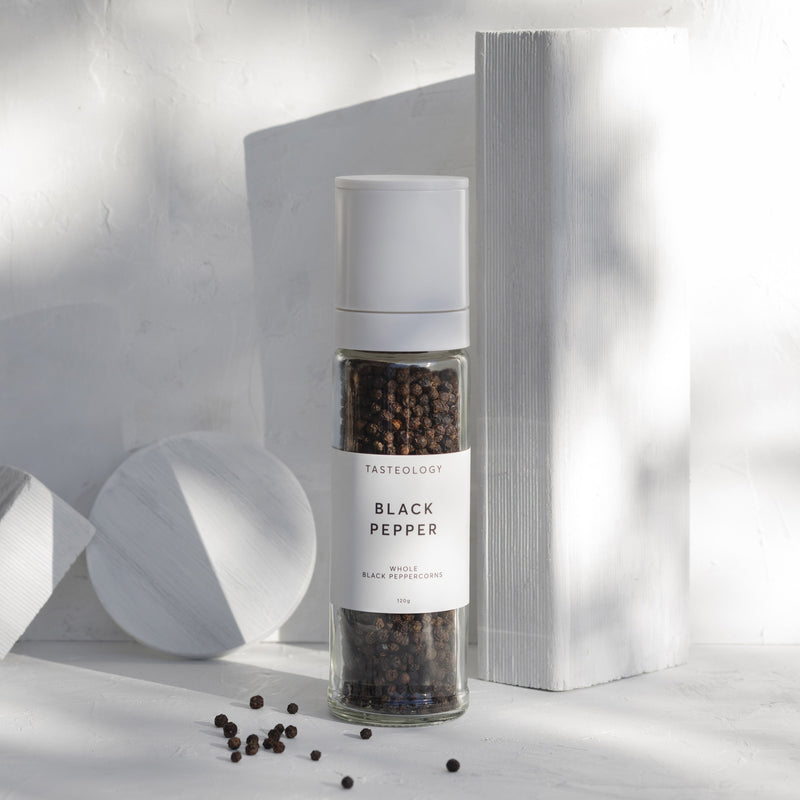 A bottle of Tasteology Black Pepper on a white surface, accompanied by a grinder.