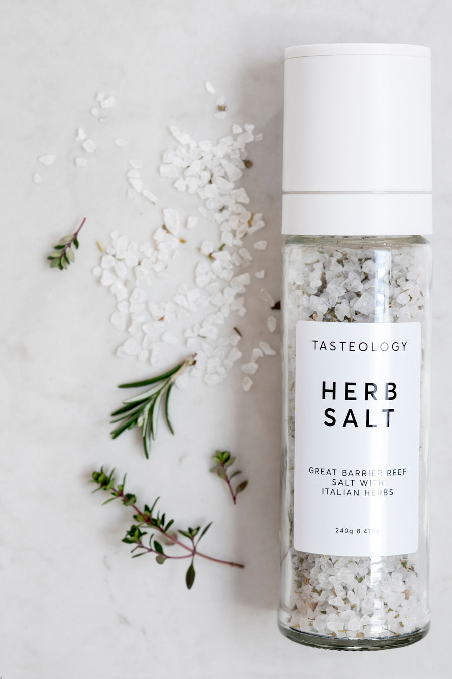 Glass bottle of Tasteology Herb Salt on marble background with scattered sea salt flakes and sprigs of rosemary and thyme.