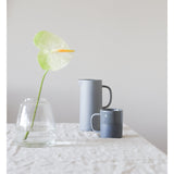 Two modern yet classic Zakkia silhouette mugs and a clear individually handmade Zakkia Tapered Vase on a table.