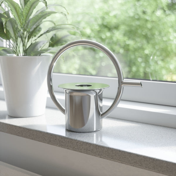 A Quench Watering Can - Stainless Steel, from the Umbra range, with a stainless steel body and a 360-degree handle, sitting on a window sill.
