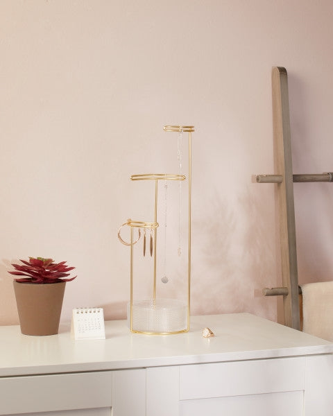 An industrial-inspired, Umbra Tesora Jewellery Stand - Glass / Brass on a white dresser next to a plant.