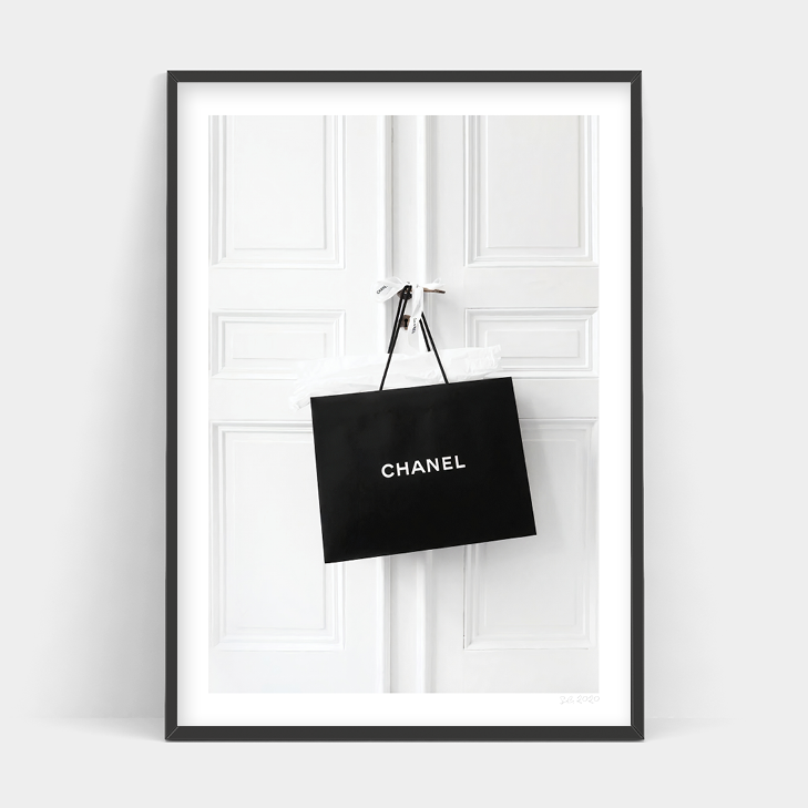 A black and white Art Prints CHANEL ADDICT shopping bag hanging on a white door.