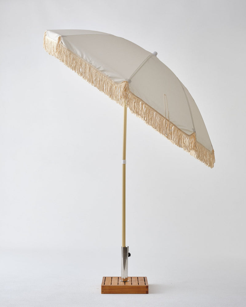 A lightweight white umbrella with tassels on a wooden stand, known for its durability, THE FRINGE UMBRELLA – PALM TREE by Brel Club.