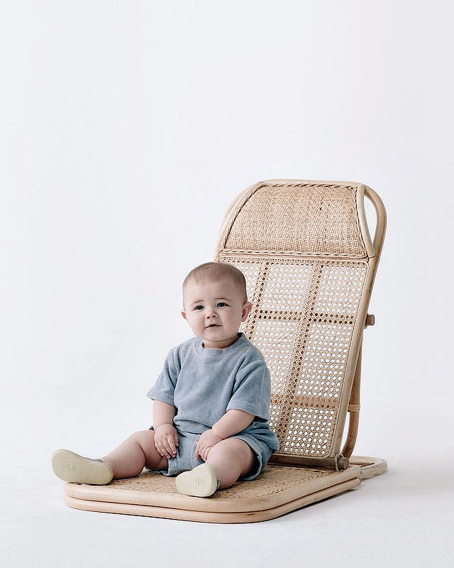 A lightweight baby sitting on THE RATTAN LOUNGER - LARGE chair by Brel Club.