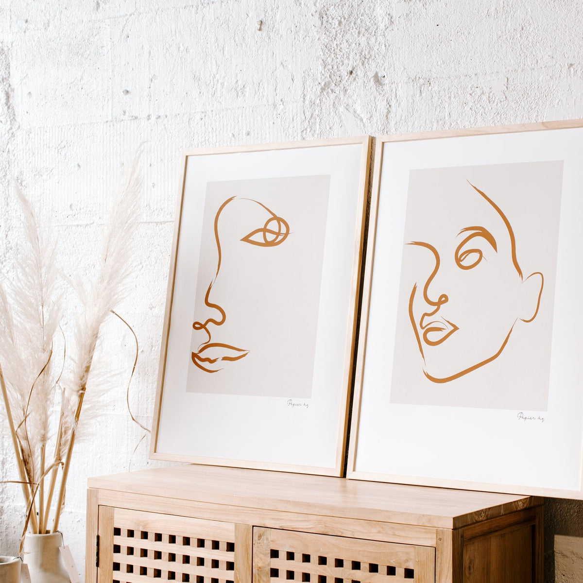 Two line face prints leaning on a cane sideboard beside dried pampas