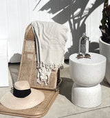 A lightweight and foldable beach lounger, THE RATTAN LOUNGER – LARGE by Brel Club, with a wicker chair, hat, and table.