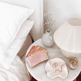 A bedside table with the Collective Hub's Affirmations to Guide Your Journey Box Card Set and a lamp on it.
