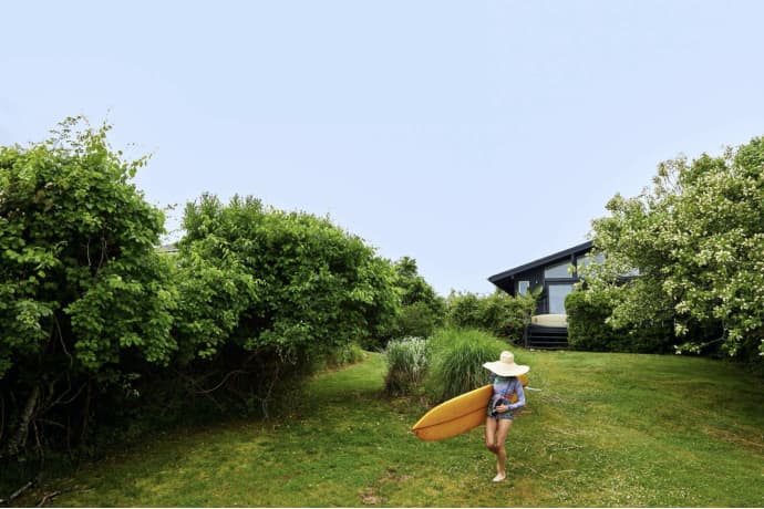 A surfer carrying a Surf Shacks Volume 2 by Gestalten in the grass near a house.
