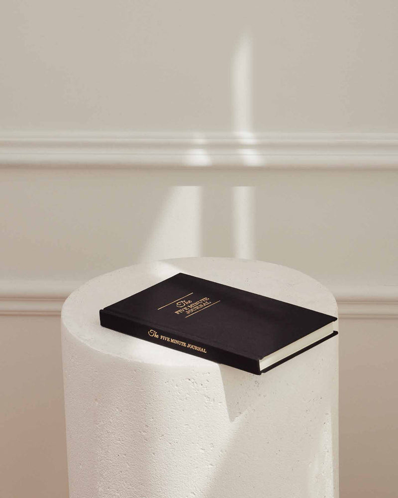 The Intelligent Change's Five Minute Journal, focused on positivity and gratitude, sits on a pedestal in front of a white wall.