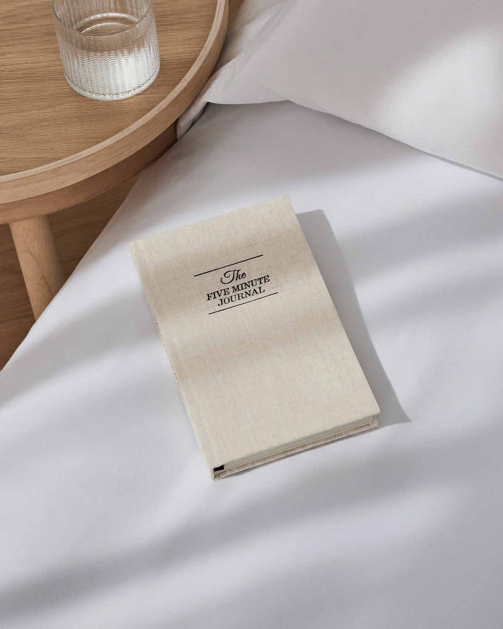 Five Minute Journal on white bedding beside a glass on a timber tray
